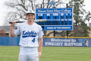 Baseball player holds bat and stands in front of scoreboard at Mount Saint Mary College. 