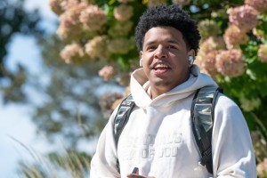 A smiling student wearing a white hoodie, wireless earbuds, and a black backpack stands outdoors among flowering trees on a sunny day.