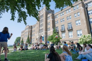 A group of students sits in a circle on a grassy lawn outside the Dominican Center, participating in an outdoor discussion beneath the shade of leafy trees on a sunny day.