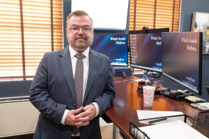 ALT: Man in a suit smiles while standing at a crowded, but organized, desk.