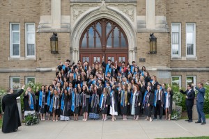 A group of graduates in caps, gowns, and blue stoles stands on steps before an ornate building entrance, celebrating with raised hands and smiles.
