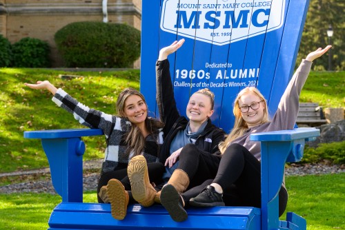 Three students sitting on a large blue Adirondack chair.