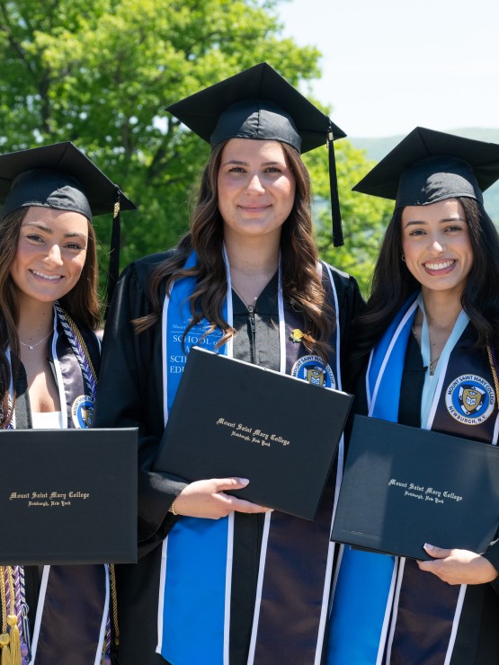 Four students stand together in graduation regalia holding their diploma case.