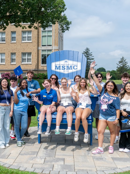 Large group of students sitting on or standing near a blue Adirondack chair on a sunny summer day.