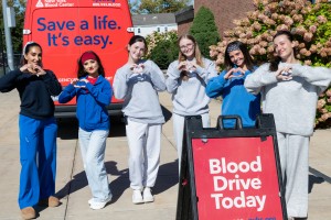 ALT: Six students making hand hearts outside in front of a red New York Blood Center van and a "Blood Drive Today" sign.