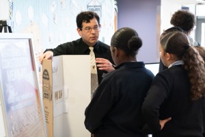 ALT: An educator speaks to school students standing around a table display. A framed sign reading "What Does the Constitution Mean to You?" rests in the foreground next to a tri-fold board featuring historical documents.