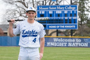 Baseball player stands holding bat. Mount Saint Mary College Business student and Knights outfielder Danny Mack stands before the scoreboard at the college's home field. 