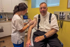 ALT: A Mount Saint Mary College nursing student, female, dressed in white and blue scrubs, gives a shot to a smiling man with glasses. 