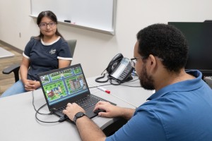 Mount Saint Mary College SURE students Ennis Cruz of Newburgh, N.Y. and Leandro Ortiz of the Bronx. ALT: A young man sits at an office desk working on a laptop displaying a top-down 2D pixel-art video game of a neighborhood, while a young woman wearing gl
