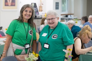 ALT: Two women smile at the camera, while festivities occur behind them.