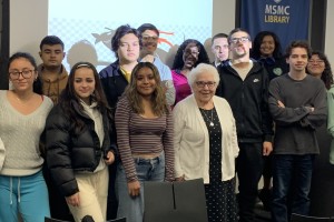 A group of college students smiling and posing around a nun in front of a projection screen.