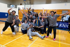 A large, excited group of college students in matching gray shirts cheering and celebrating around a tall blue trophy on a basketball court.