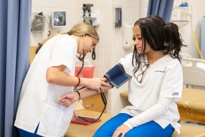 ALT: A nursing student wearing white scrubs uses a stethoscope and blood pressure cuff to check the blood pressure of another student in a clinical lab setting.