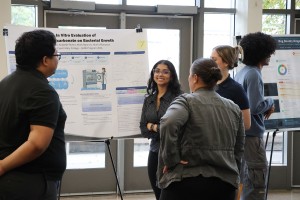 ALT: A smiling student researcher with glasses stands next to her scientific poster on bacterial growth while discussing it with a small group of attendees.