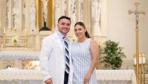A smiling couple stands in a church near the altar. The man wears a white coat and striped tie; the woman wears a striped dress. The background features religious statues and candles, conveying a celebratory and serene atmosphere.