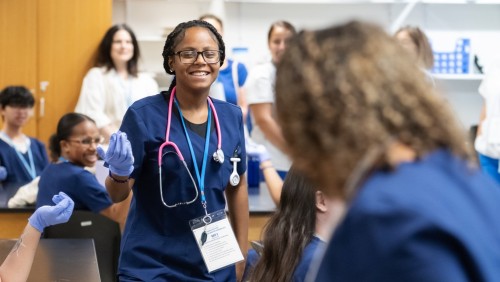 A smiling person in scrubs and a stethoscope stands in a classroom, exuding confidence. Surrounded by peers in similar attire, the atmosphere is lively and engaging.