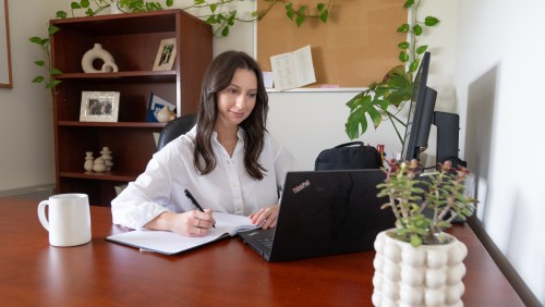 An adult student in a white shirt working at a desk with a laptop, notebook, and pen. Shelves, plants, and a corkboard are in the background. 