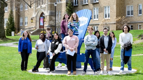 Group of students standing on or near a large Adirondack chair.