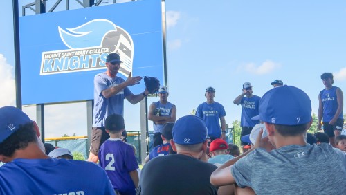 A group of young baseball players sits attentively on the grass, listening to a coach speaking energetically. A large "Mount Saint Mary College Knights" sign is visible in the background, creating an inspiring and educational atmosphere.
