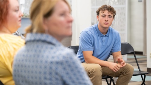 Graduate student in a blue polo shift attentively listens in a group discussion. Others seated around him are blurred.