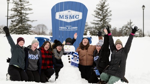 Seven students in snow gear posing with a snowman in a Mount Saint Mary College scarf with a large Adirondack chair behind them.