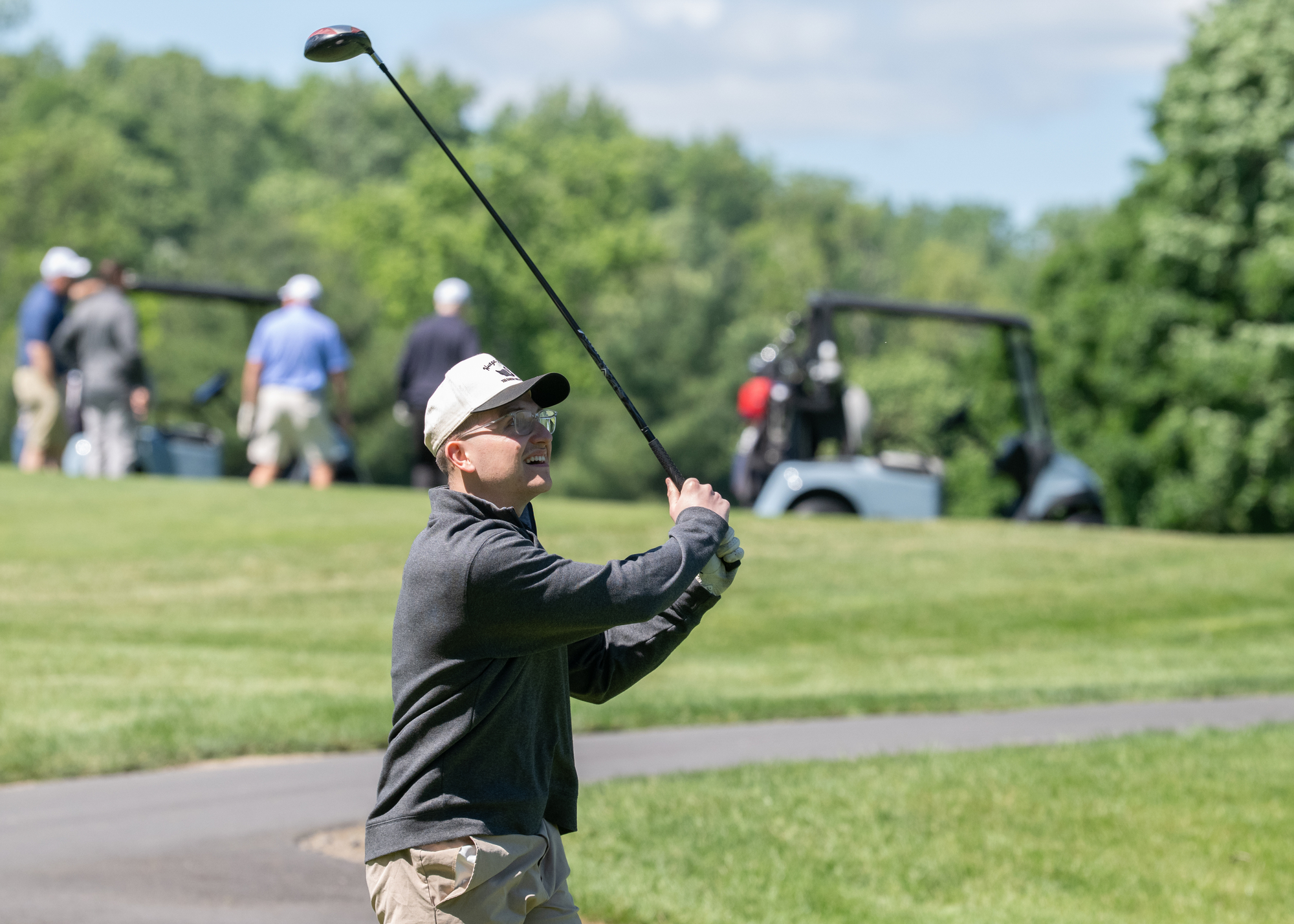 A golfer in a cap and sunglasses swings a club on a sunny course, while others and golf carts are visible in the background amid lush greenery.