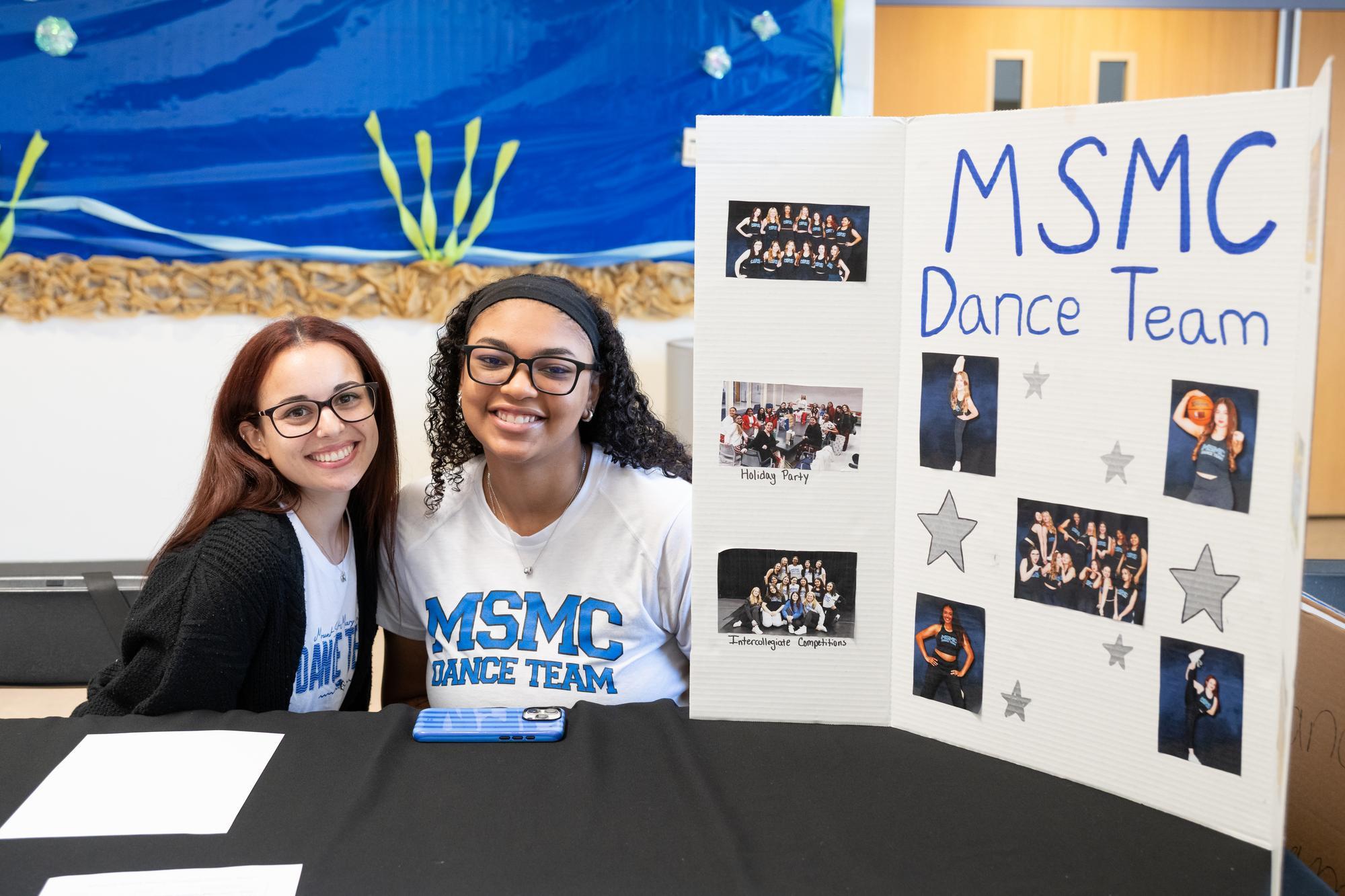 Two smiling students sit at a table promoting the MSMC Dance Team. A poster with photos and text is displayed.