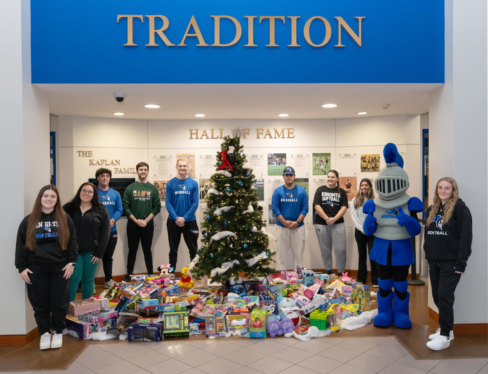 A group of students with the campus mascot stand smiling around a decorated Christmas tree surrounded by donated toys, under a "Tradition" sign and in front of the Hall of Fame.